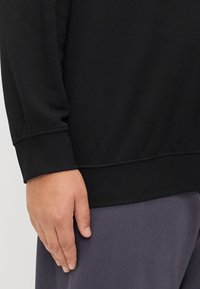 Sudadera negra con una textura suave, puños acanalados y un corte relajado. Mano descansando sobre un pantalón gris, mostrando colores de tejido contrastantes.