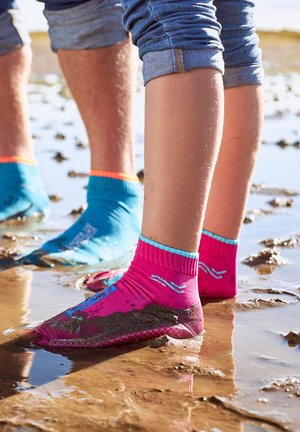 Zwei Personen tragen bunte Wasserschuhe und hochgekrempelte Jeans, während sie in flachem schlammigem Wasser im Freien stehen.