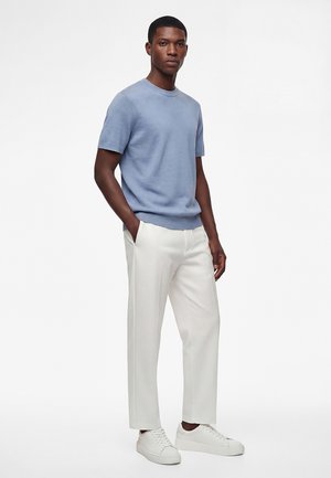 Man draagt lichtblauw gebreid shirt met korte mouwen, witte op maat gemaakte broek en witte sneakers, staat met handen in de zakken op een witte achtergrond.