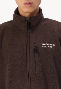 Brązowa polarowa kurtka z wysokim kołnierzem. Posiada zapinane na zamek kieszenie oraz biały logo "GAP/SPORT EST. 1969" z przodu. Miękka tekstura.