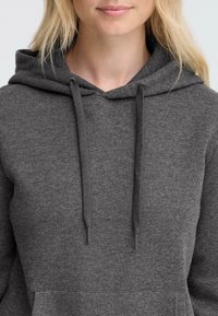 Personne blonde portant un sweat à capuche gris foncé avec cordons, visible des épaules à la bouche, fond neutre.