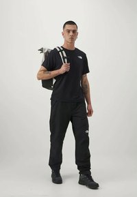 Schwarzes T-Shirt und Hose mit einem Logo. Das Modell trägt einen Rucksack mit grauen Akzenten. Das Material wirkt strapazierfähig und hat eine lässige Passform.