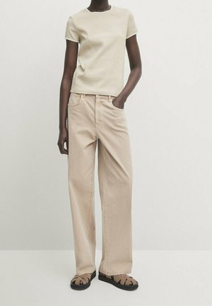 Slim dark-skinned person wearing a beige short-sleeve top, loose beige pants, and brown sandals, standing with one hand in pocket.