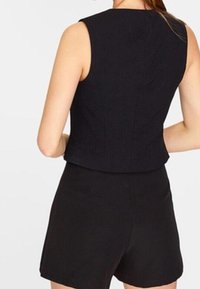 Kvinna i en ärmlös svart topp och matchande svarta högmidjade shorts, sedd bakifrån mot en neutral bakgrund.