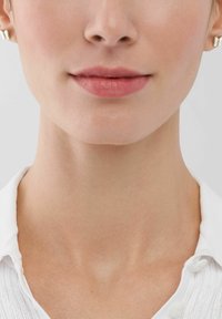 Orecchini a bottone in oro su una carnagione chiara, con un colore naturale per le labbra. Il soggetto indossa una camicia bianca con colletto e un motivo testurizzato.