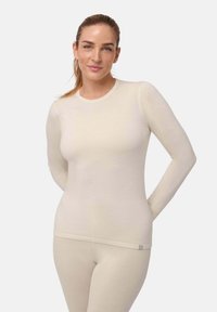 Kobieta z jasnobrązowymi włosami związanymi z tyłu, ubrana w dopasowaną kremową bluzkę z długim rękawem i pasujące spodnie, stoi z rękami za plecami.