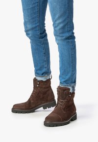 Bottines en daim brun avec un bout arrondi, un système de laçage à l'avant et des accents en métal. La surface texturée et la semelle noire robuste sont visibles.