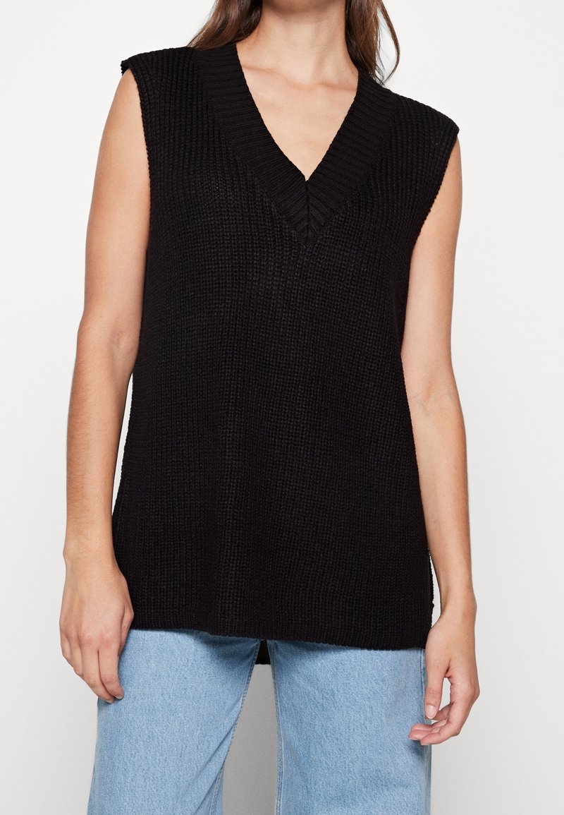 Femme portant un gilet sans manches en tricot noir avec col en V et un jean bleu clair, montrée des épaules aux hanches sur fond blanc.