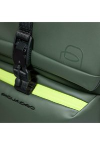 Mochila verde con acabado texturizado, que presenta una hebilla negra, un llamativo cierre de cremallera amarillo y un detalle de logotipo en relieve sobre el material.