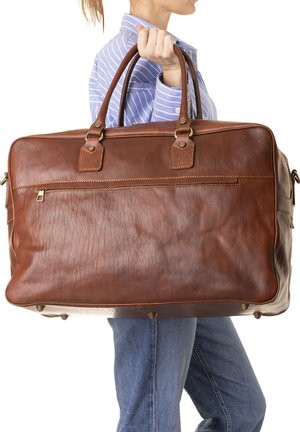 Persoon in een blauw gestreept shirt en spijkerbroek houdt een grote bruine leren reistas vast bij de handvatten.