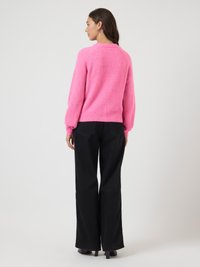 Mulher vista de costas, vestindo um suéter rosa brilhante e felpudo, calças largas pretas e sapatos de salto pretos, contra um fundo claro e simples.