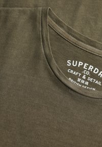 Oliwkowozielony bawełniany t-shirt z okrągłym dekoltem, z nadrukowanym białym logo: "SUPERDRY CO. CRAFT & DETAIL" na przodzie.