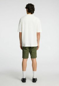 Krämfärgad t-shirt med korta ärmar och avslappnad passform, parat med olivgröna shorts. Modellen bär svarta skor och vita ankelsockor.