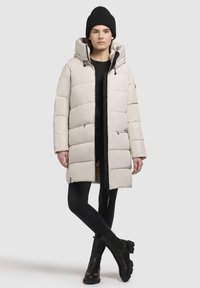 Beige gewatteerde lange jas met een ritssluiting aan de voorkant, capuchon en elastische bindings. Draagt over zwarte leggings en zwarte enkellaarsjes.