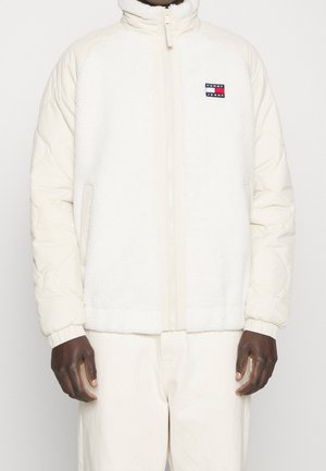 Osoba nosząca białą kurtkę polarową z pikowanymi rękawami i logo Tommy Jeans na piersi, w połączeniu z białymi spodniami.