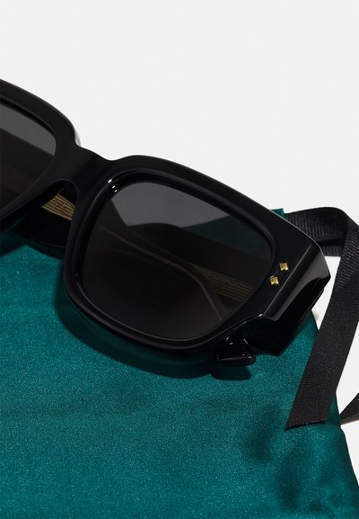 Black rectangular sunglasses with dark lenses, featuring gold accents on the frame, resting on a textured teal fabric pouch.