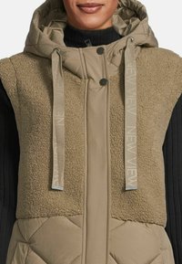 Woman wearing a beige hooded vest with quilted and fleece textures and drawstrings labeled "NEW VIEW" over a black ribbed sweater.