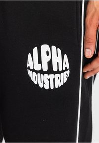Schwarze Jogginghosen mit einem weißen "ALPHA INDUSTRIES"-Logo. Aus Baumwollmaterial mit einer weichen Textur und kontrastierenden weißen Seitenstreifen. Nahaufnahme.