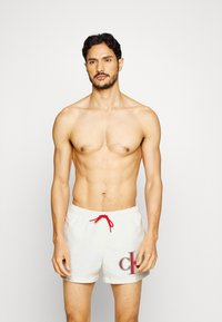 Costumi da bagno da uomo in beige chiaro con un laccio rosso. Presentano un grande logo nero "CK" sulla coscia sinistra. Tessuto liscio e vestibilità comoda.