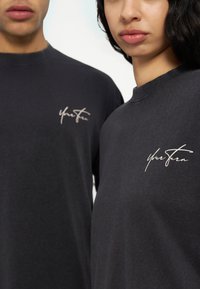 Dos personas con camisetas gris oscuro con el texto "Tu turno" bordado en el pecho, primer plano de torsos superiores y rostros.