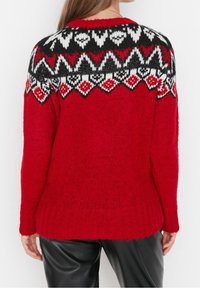 Rood gebreid sweater met een zwart, wit en rood geometrisch patroon op de bovenrug en schouders, gedragen met zwarte broek.