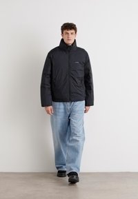 Zwarte pufferjack met een hoge kraag, ritssluiting en voorzak, gedragen over lichtblauwe jeans en zwarte schoenen. Effen achtergrond.