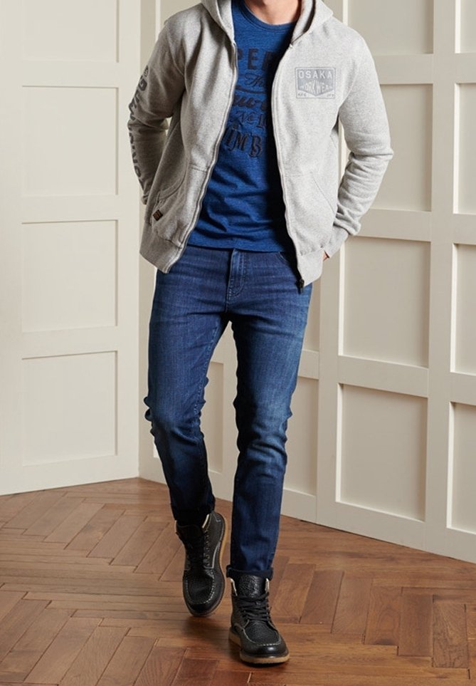 Mand i en grå hættetrøje med lynlås, blå grafisk t-shirt, slanke blå jeans og sorte høj-top sneakers, stående på trægulv.