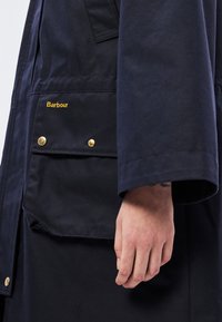 Αδιάβροχο μπουφάν σε ναυτικό μπλε χρώμα με χρυσές λεπτομέρειες, λογότυπο στην τσέπη και χαλαρή εφαρμογή με διακριτές ραφές.