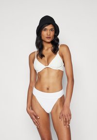 Bílý žebrovaný bikinový set s trojúhelníkovým braletem a vysokým pasem. Obsahuje tenké ramínka a hladkou texturu. Nosený s černým pleteným kloboukem.
