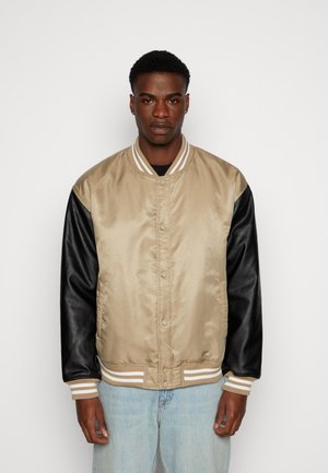 Beige Satin-Bomberjacke mit schwarzen Lederärmeln, gestreiftem Rippkragen und Druckknopfverschluss, kombiniert mit hellblauen Jeans.