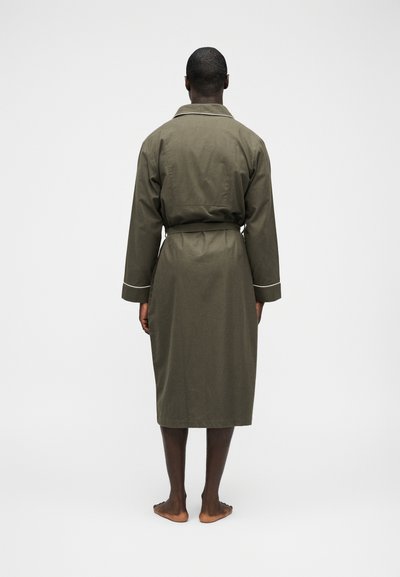 Άνδρας που στέκεται ξυπόλητος με την πλάτη γυρισμένη, φορώντας μια ρόμπα μήκους έως το γόνατο σε ελιά πράσινο χρώμα με λευκές ρίγες στα μανίκια και στον γιακά, μπροστά σε απλό φόντο.