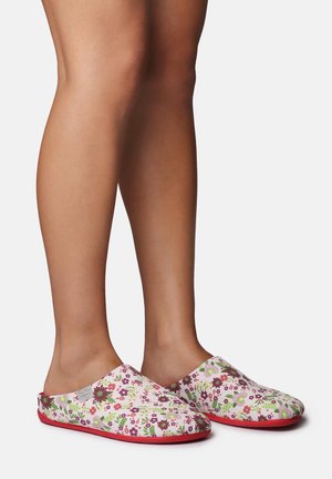 Nohy v bielych slip-on papučiach s červenými podrážkami, pokryté viacfarebným kvetinovým vzorom, stojace na jednoduchom pozadí.