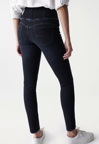 Osoba v tmavých úzkých džínech s vysokým pasem a bílých teniskách, stojící s jednou rukou uvolněně podél těla.