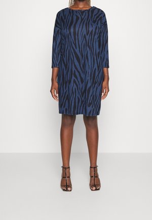 Vrouw die een blauw-zwart jurk met zebraprint tot op de knie draagt, met driekwartmouwen en zwarte sandalen met hoge hakken en bandjes.