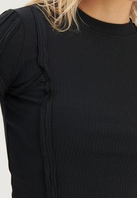 Schwarzes geripptes Top mit einem runden Ausschnitt und strukturiertem Rüschendetail entlang der Seitennaht für zusätzlichen visuellen Reiz.