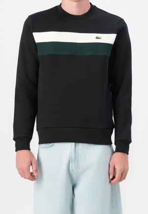 Mężczyzna w czarnej bluzie z białymi i ciemnozielonymi poziomymi paskami na wysokości klatki piersiowej oraz jasnoniebieskich dżinsach.