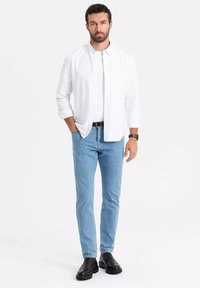 Muž stojící s rukama, oblečený v bílém košili přes bílém tričku, světlých modrých džínách, černém opasku, černých botách a s hodinkami na bílém pozadí.