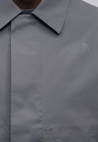 Γκρι εξωτερικό μπουφάν με δομημένο γιακά, διαθέτει ανάγλυφο λογότυπο στην πάνω αριστερή πλευρά και λείο φινίρισμα υφάσματος.