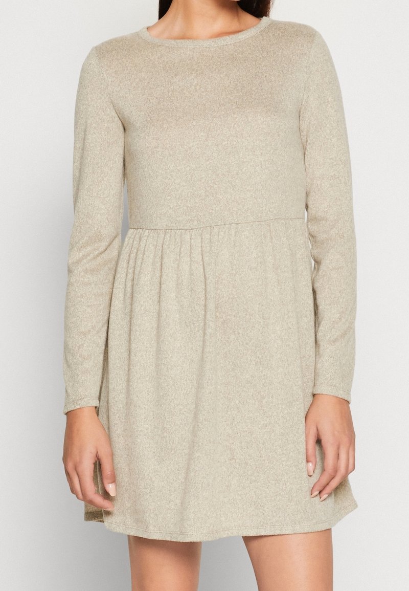Femme portant une robe beige à manches longues avec un corsage ajusté et une taille froncée, debout devant un fond uni.
