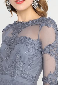 Grå klänning med spetsbroderi längs halsringningen och ärmarna. Transparent meshmaterial har intrikata mönster och texturerade accenter.