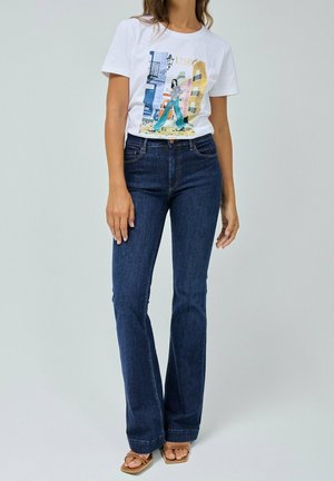 Kobieta ubrana w białą koszulkę z nadrukiem miasta Lizbona, ciemnoniebieskie dżinsy bootcut oraz beżowe sandały z odkrytymi palcami, stojąca na jednolitym tle.