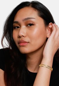 Bracciale in oro con anelli intrecciati, indossato su un polso, con una finitura lucida su una camicia nera dal tessuto morbido e liscio.