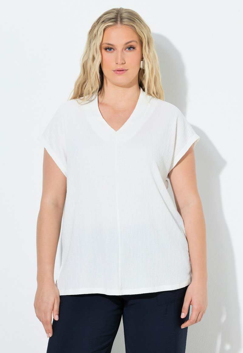 Femme blonde aux cheveux ondulés, portant un haut blanc à manches courtes en V et un pantalon foncé, debout devant un fond blanc uni.