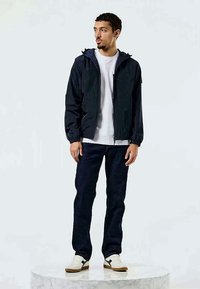Casaco corta-vento azul-marinho com fecho de zip, punhos elásticos e capuz ajustável, vestido sobre uma t-shirt branca e jeans escuros, com sapatilhas neutras.