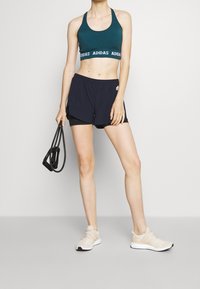 Pantalones cortos deportivos de color azul oscuro con pantalones cortos negros por debajo, sujetador deportivo en color verde azulado con la marca blanca "ADIDAS" y zapatillas de correr en color beige.