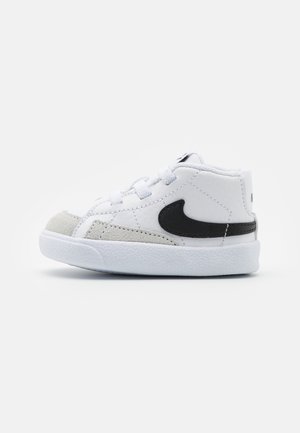 Sneaker pour tout-petit blanc avec le logo Nike noir, lacets blancs, détails en daim gris sur la pointe et semelle blanche texturée sur fond uni.