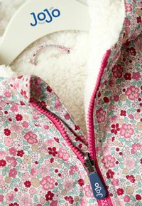 Jachetă pentru bebeluși cu fermoar, în culori roz și roșu cu model floral, căptușită cu fleece alb pufoș, agățată pe un suport alb Jojo.