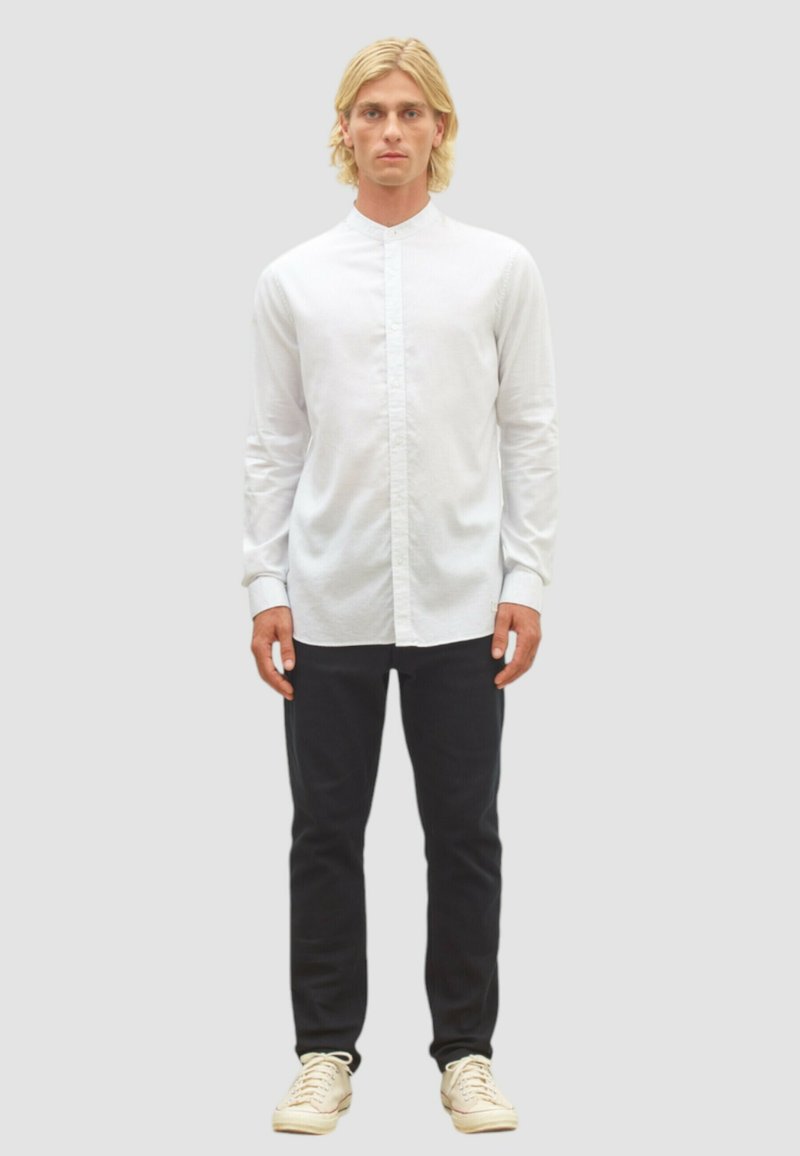 Weißes Hemd mit Stehkragen, kombiniert mit dunklen Hosen und hellen Sneakers, vor einem neutralen Hintergrund.