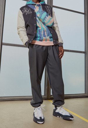 Schwarz-cremefarbige Varsity-Jacke, mehrfarbige Kapuzenjacke, graue Hose, weiße und schwarze Sneaker sowie eine blaue Umhängetasche mit Logo-Akzenten.