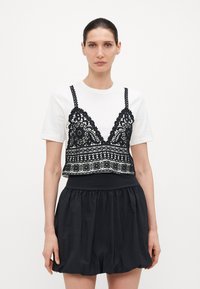 Čierny čipkový crop top s zložitými vzormi na bielom tričku, doplnený o čiernu, nariasenú sukňu. Všetky kusy sú z ľahkej látky.